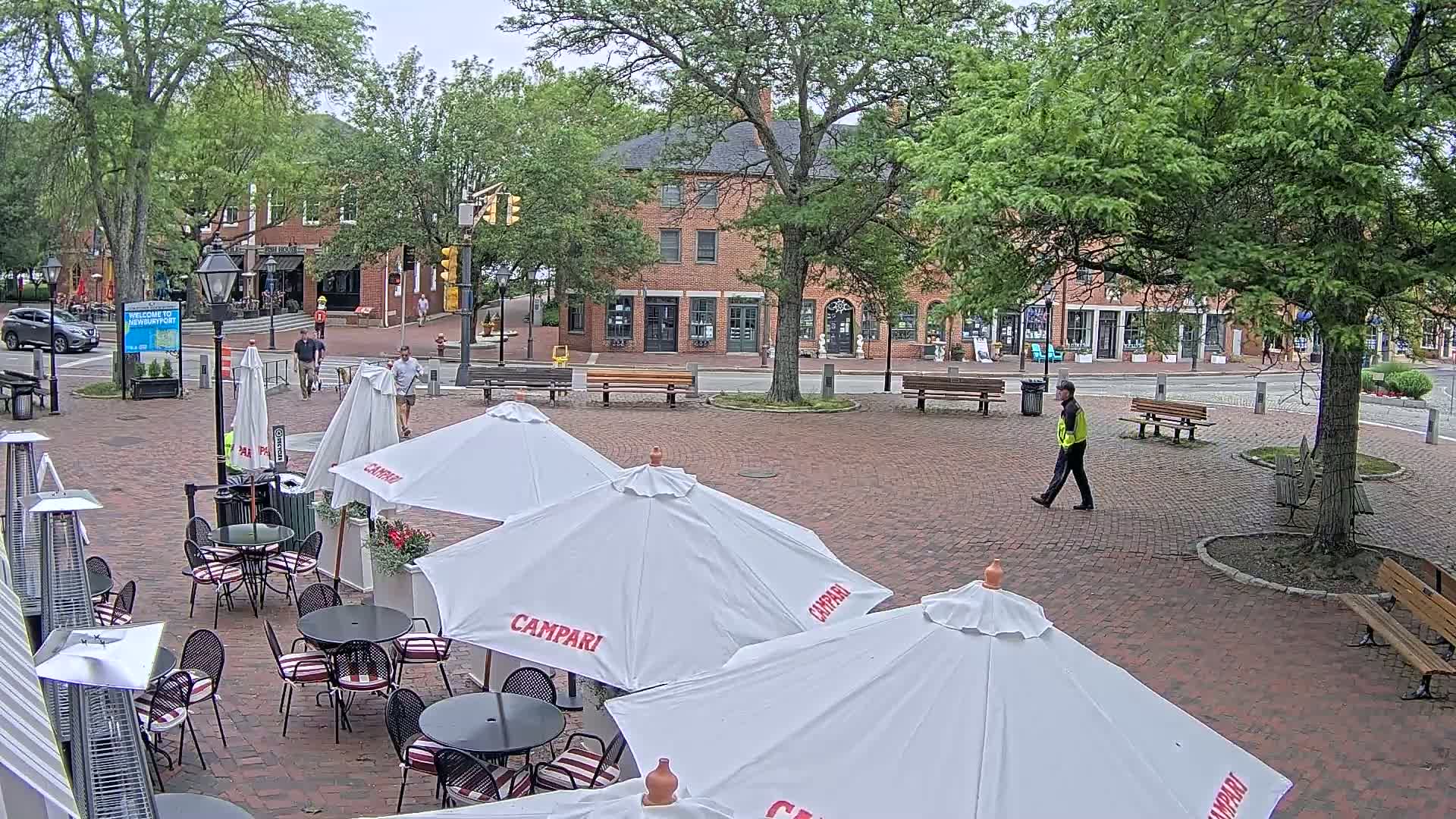 A brick plaza with several patio umbrellas and outdoor seating, surrounded by trees and brick buildings, on an overcast day.
