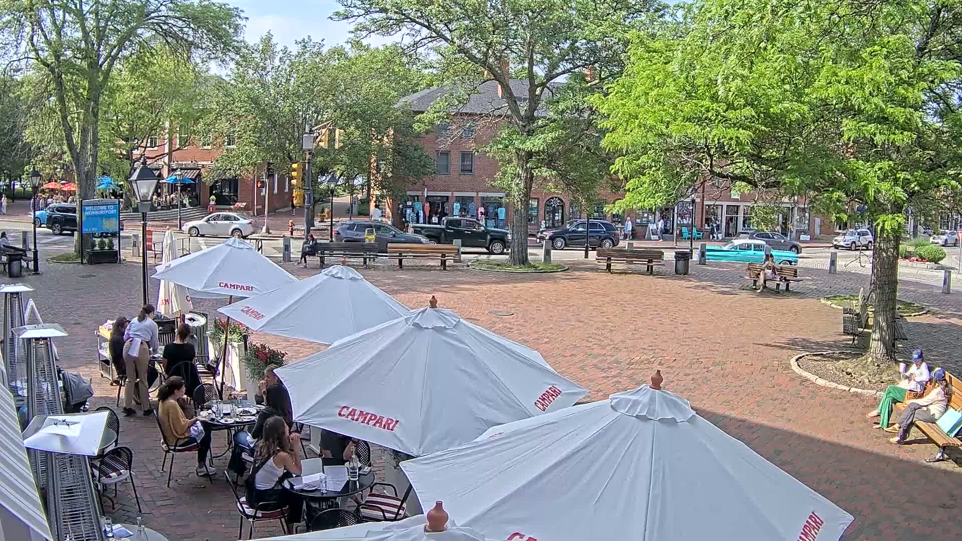 An outdoor brick plaza with several cafe umbrellas, patrons seated at tables, and surrounding buildings under a sunny sky.