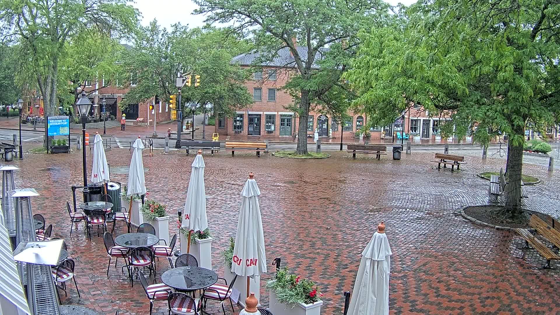 A wet brick plaza with outdoor cafe seating under white umbrellas, surrounded by brick buildings and lush green trees in the rain.