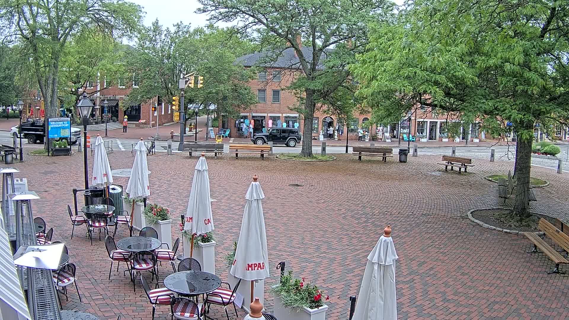 A brick town square with several outdoor cafe umbrellas, benches, and a parked Jeep, under an overcast sky.