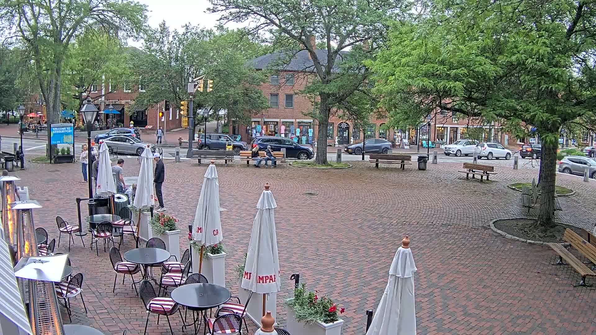 A brick town square with several outdoor cafe umbrellas, benches, and a parked Jeep, under an overcast sky.