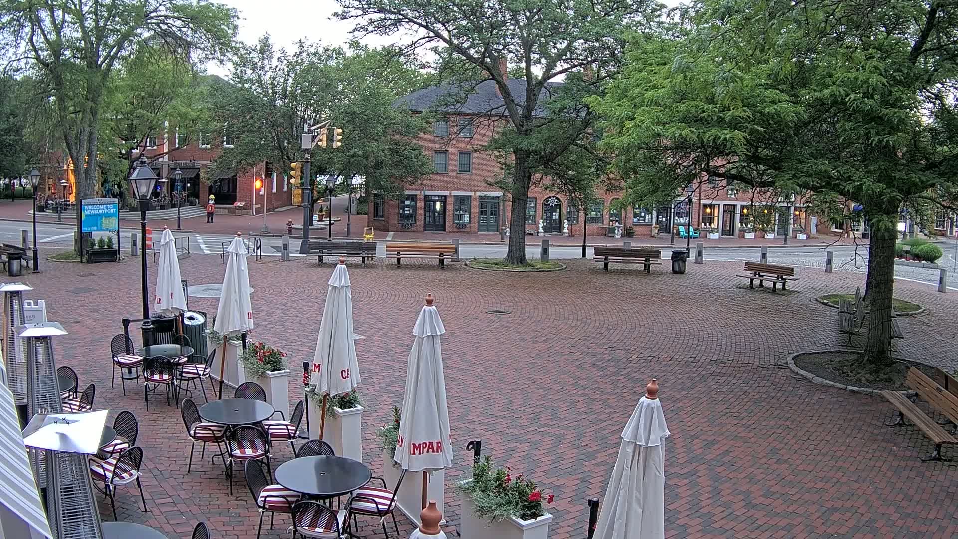 A brick plaza with outdoor café seating under white umbrellas, surrounded by brick buildings and lush green trees under an overcast sky.