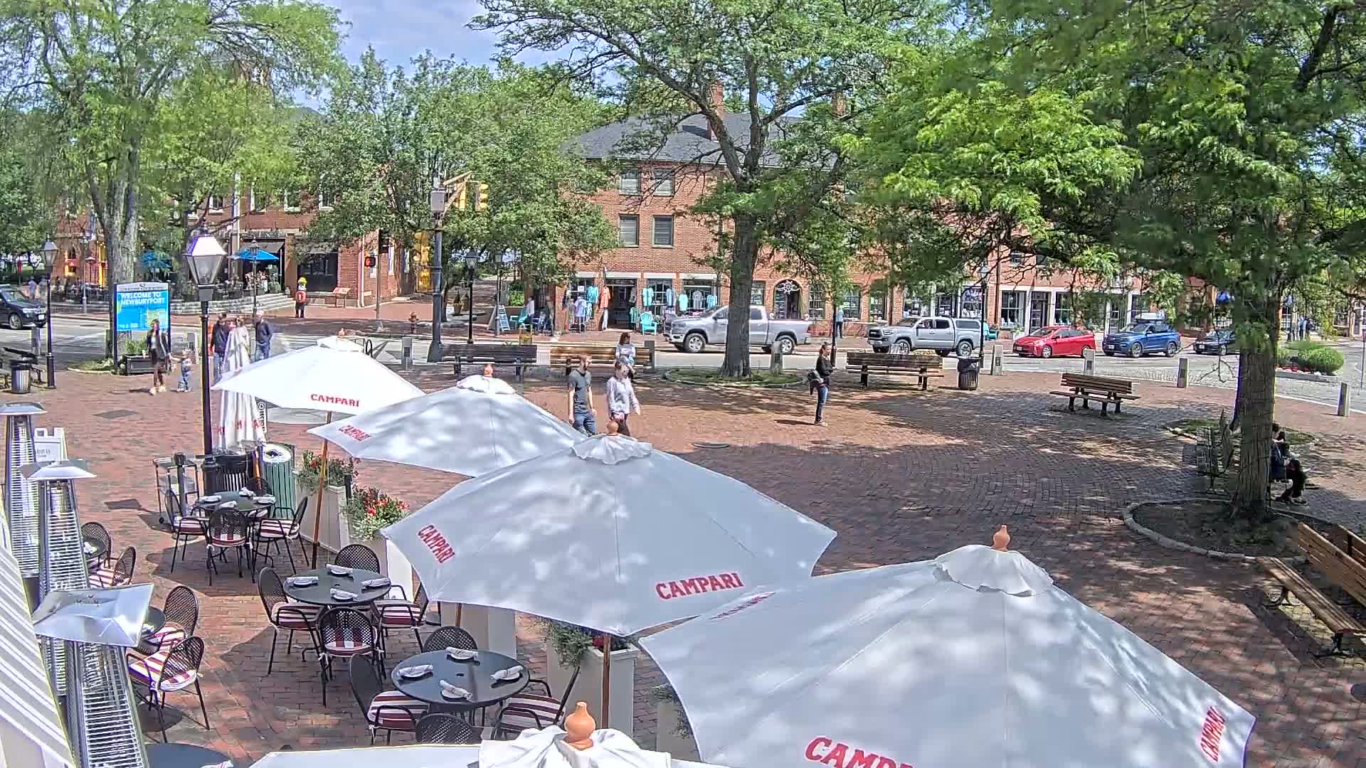 A brick plaza with several outdoor cafe umbrellas, surrounded by brick buildings and lush green trees under a partly sunny sky.