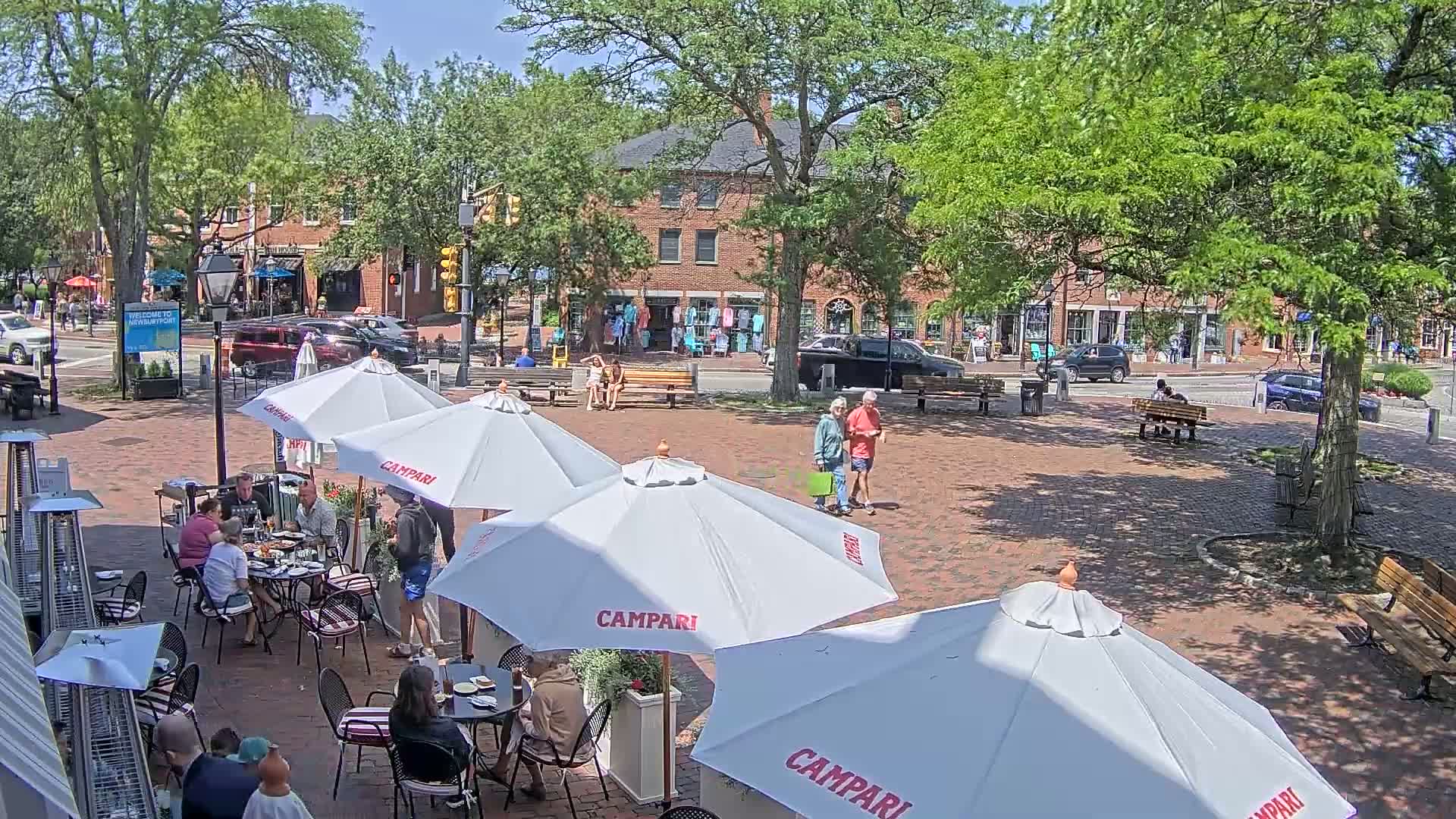 A brick plaza with several outdoor cafe seating areas under white umbrellas, surrounded by brick buildings and lush green trees under a sunny sky.