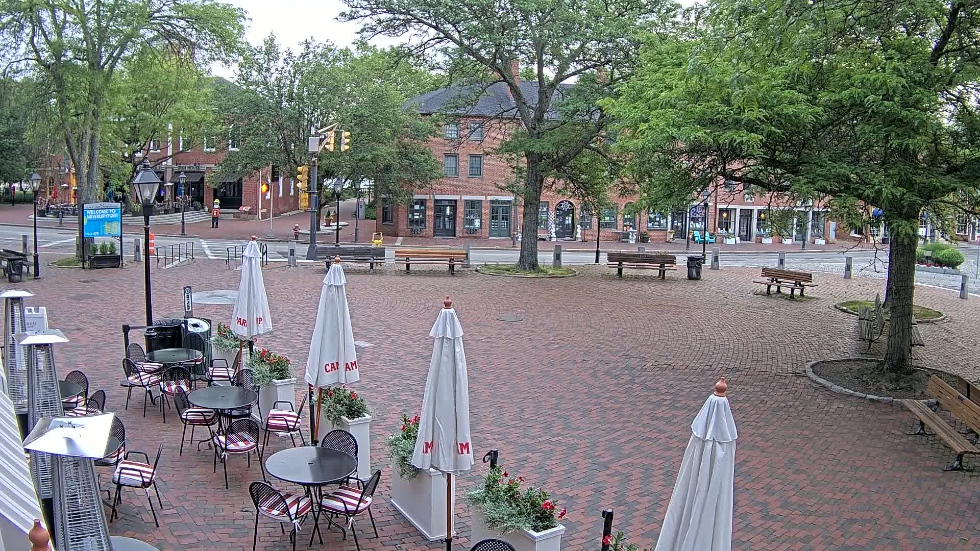 An outdoor brick patio with cafe tables and umbrellas is situated in a town square, with brick buildings and lush green trees under an overcast sky.