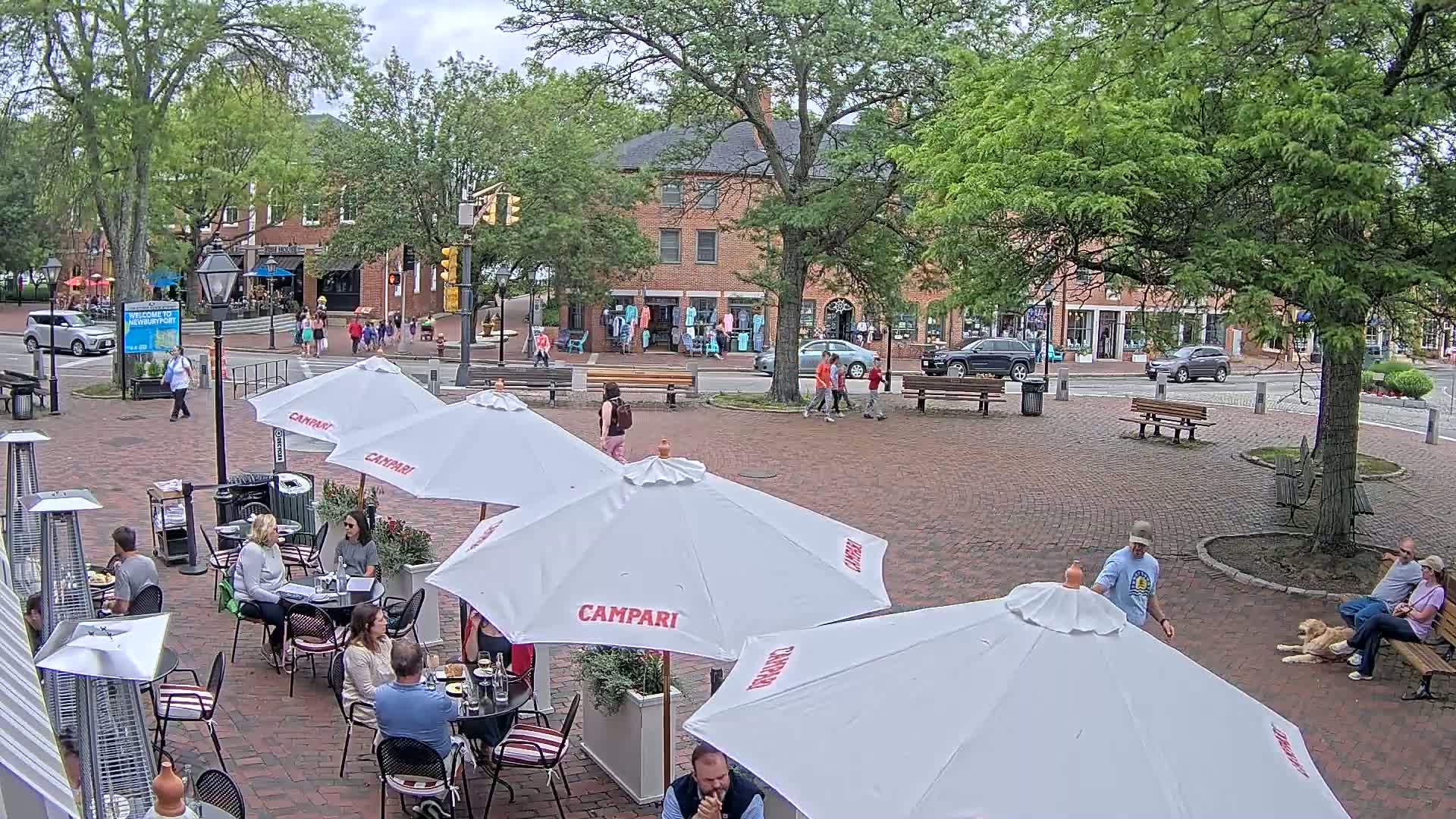 An outdoor brick plaza with several patio umbrellas and diners, under a partly cloudy sky, is surrounded by shops and trees.