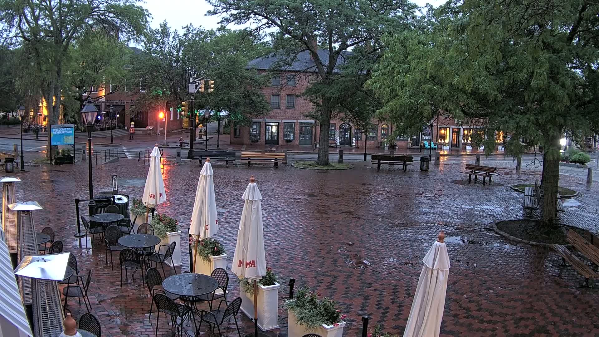 A wet cobblestone town square with several patio umbrellas and tables, surrounded by brick buildings and trees under an overcast sky.