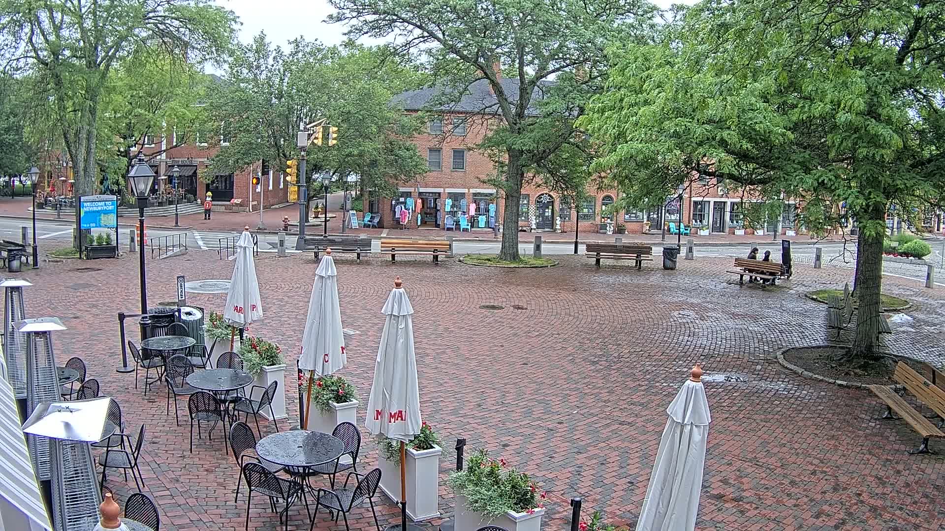 A brick plaza with outdoor seating under white umbrellas, a few people sitting on a park bench, and brick buildings in the background on an overcast day.