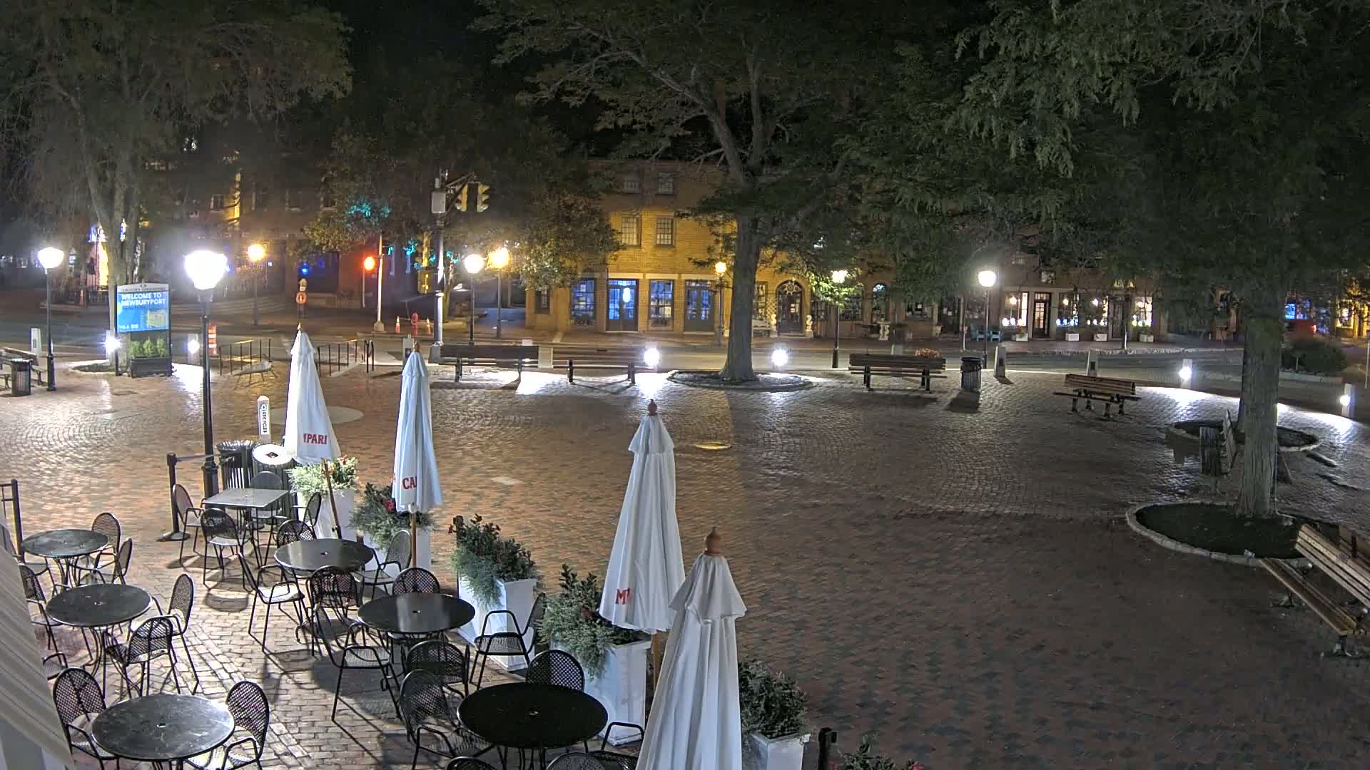 A brick plaza at night, lit by streetlights, contains several bistro tables with white umbrellas, benches, and a large tree in the center, surrounded by buildings.