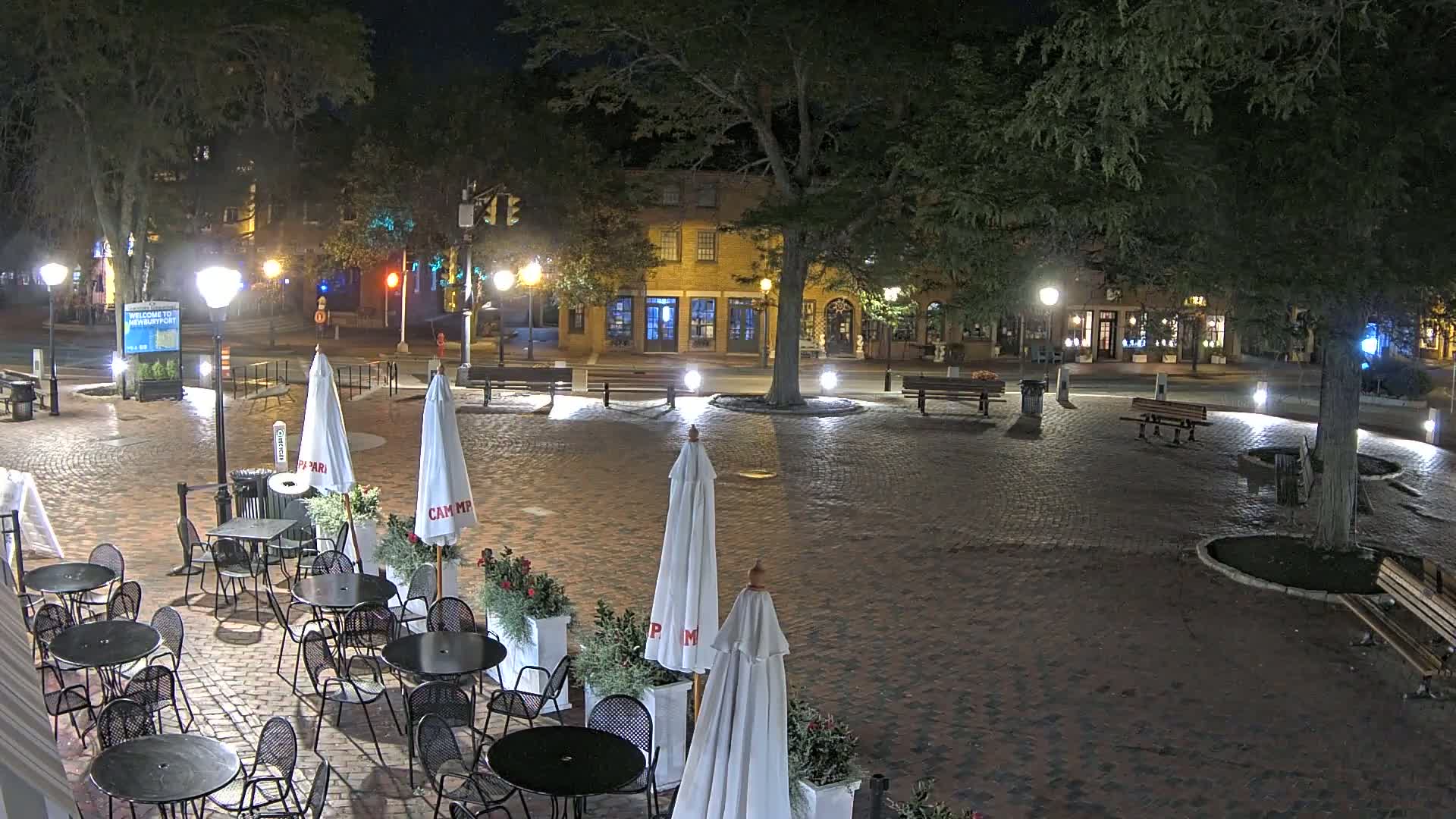 A brick plaza at night, illuminated by lampposts, with outdoor seating, trees, and buildings visible in the background.