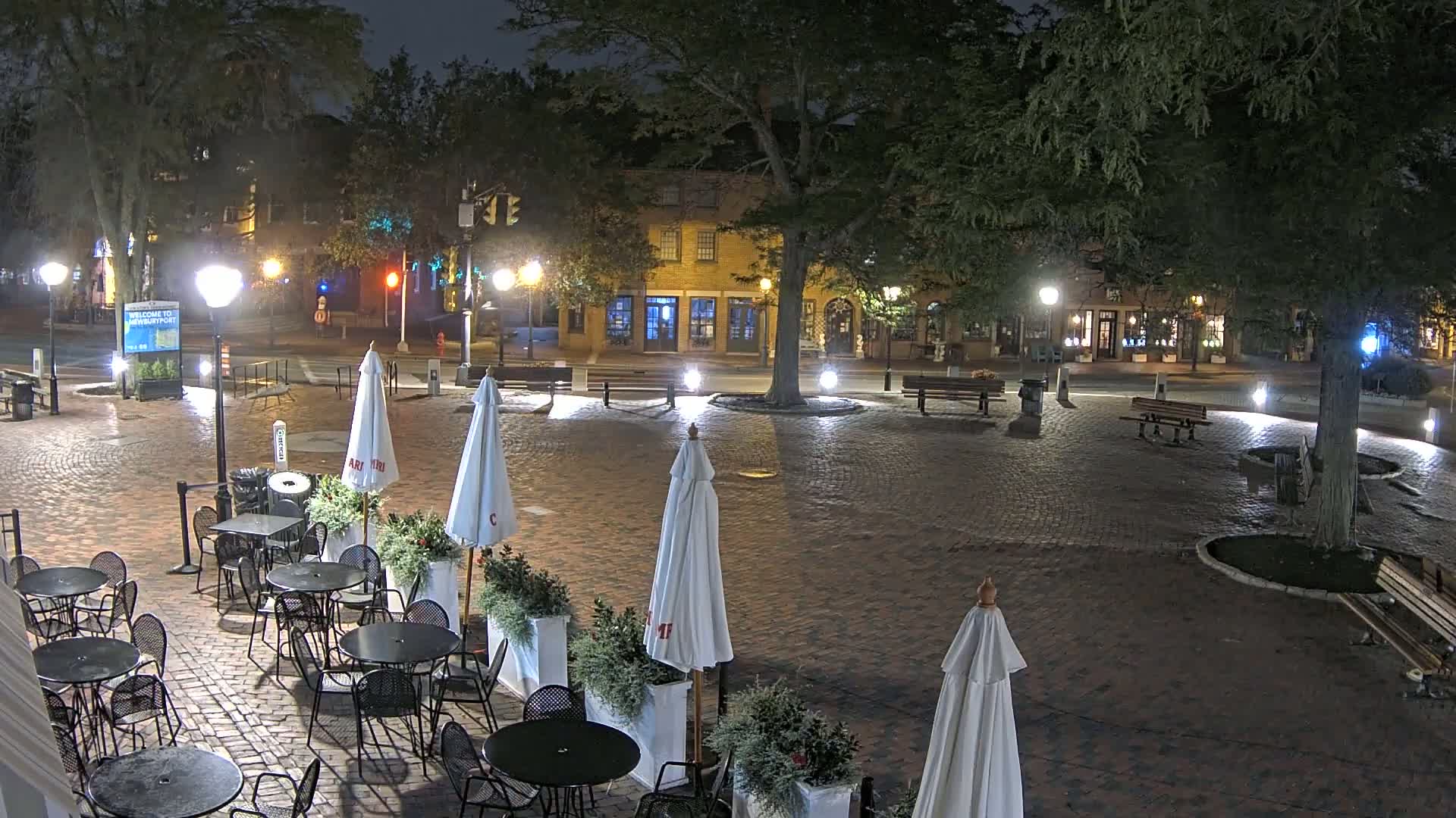 A brick plaza at night, illuminated by lampposts, with several outdoor tables and umbrellas set up around a central tree, and a street with buildings visible in the background.