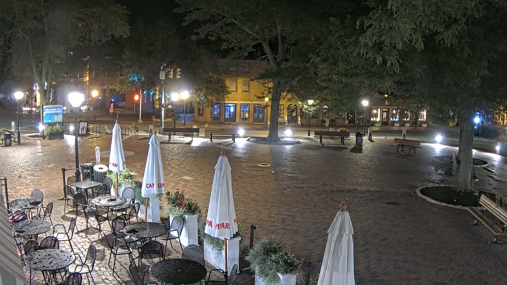 A brick plaza at night, illuminated by streetlights, with outdoor café seating, potted plants, and trees surrounding the area.