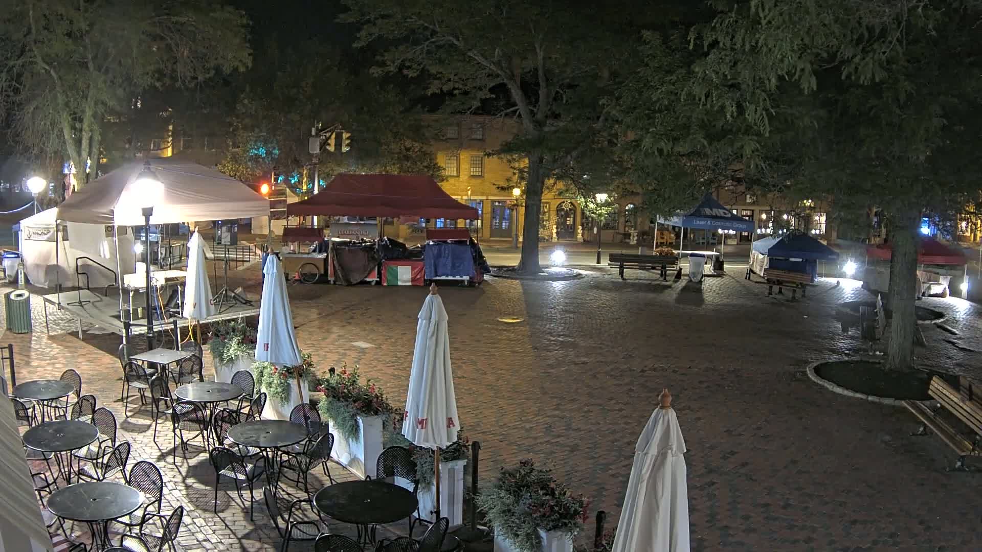 A brick plaza at night, illuminated by lampposts, features several market stalls under canopies, outdoor seating with umbrellas, and trees.