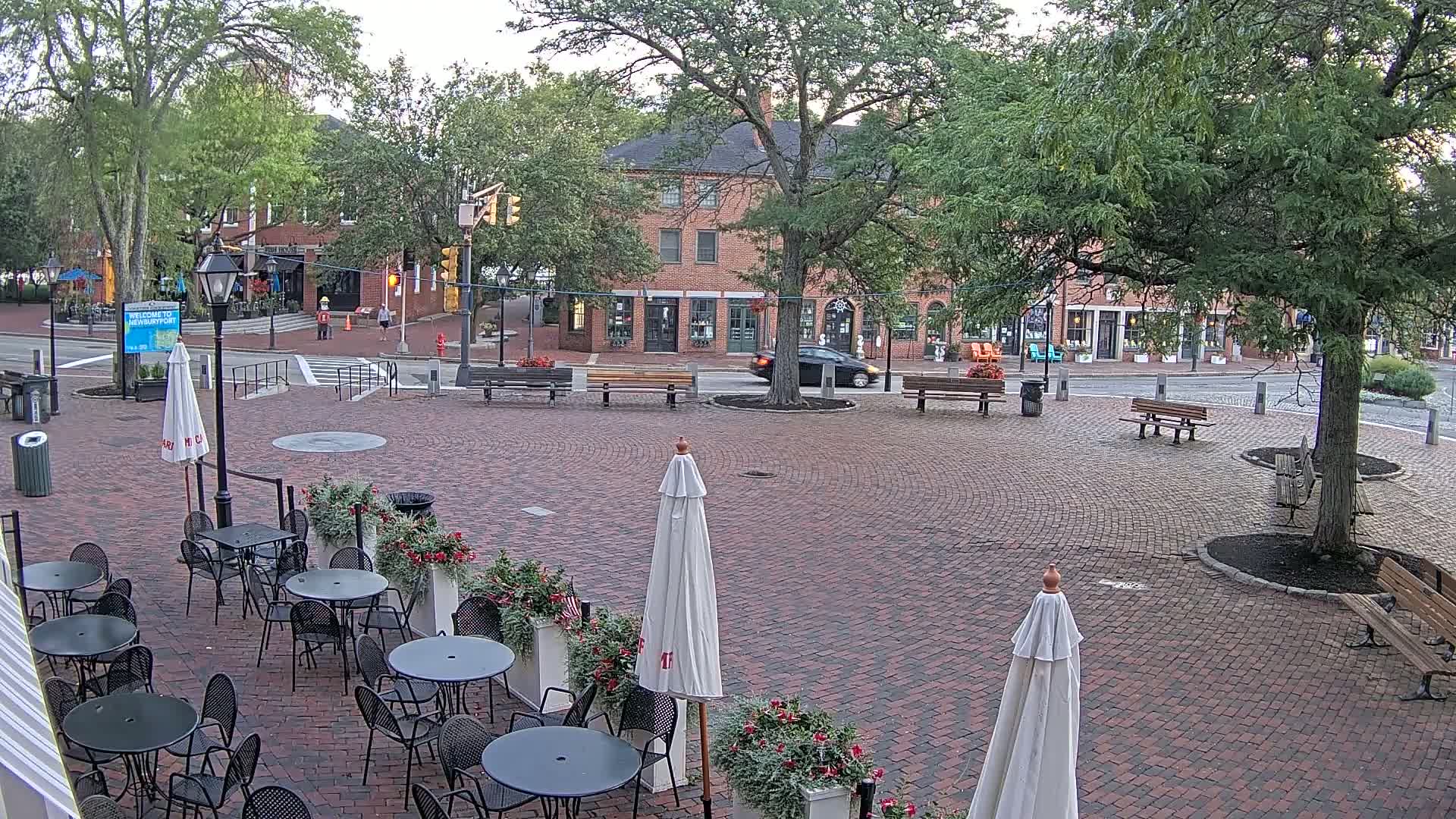 A brick plaza at night, illuminated by lampposts, features several market stalls under canopies, outdoor seating with umbrellas, and trees.