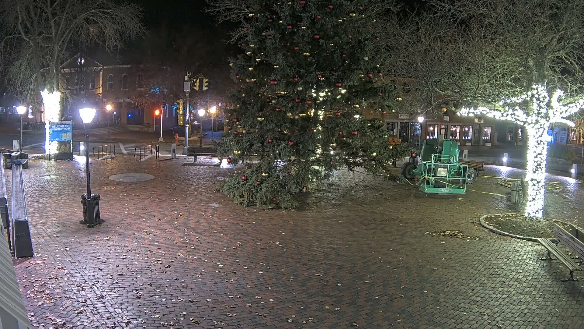 A festive town square is seen at night under clear skies, featuring a large, decorated Christmas tree, a tree wrapped in white lights, and a green hydraulic lift parked on brick pavement scattered with leaves.