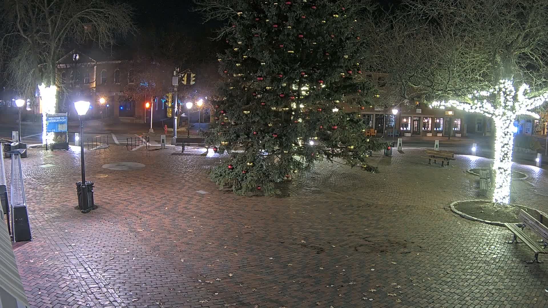 An empty brick-paved town square is illuminated at night by a large decorated Christmas tree and a bare tree wrapped in white lights under clear skies.
