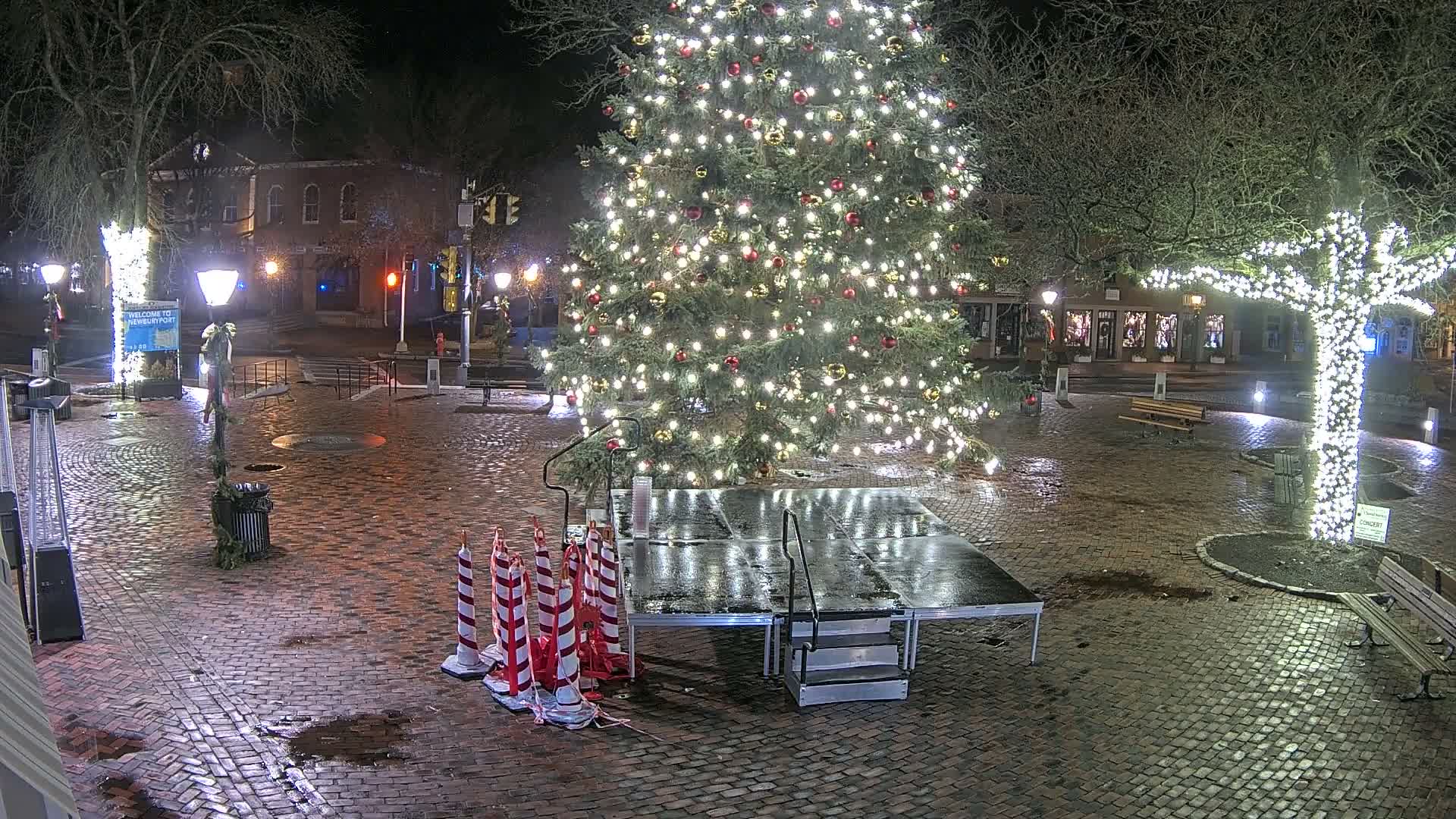 A festive town square is seen at night under damp conditions, featuring a large, brightly decorated Christmas tree, a small stage, and trees wrapped in white lights, all illuminating the wet cobblestone ground.