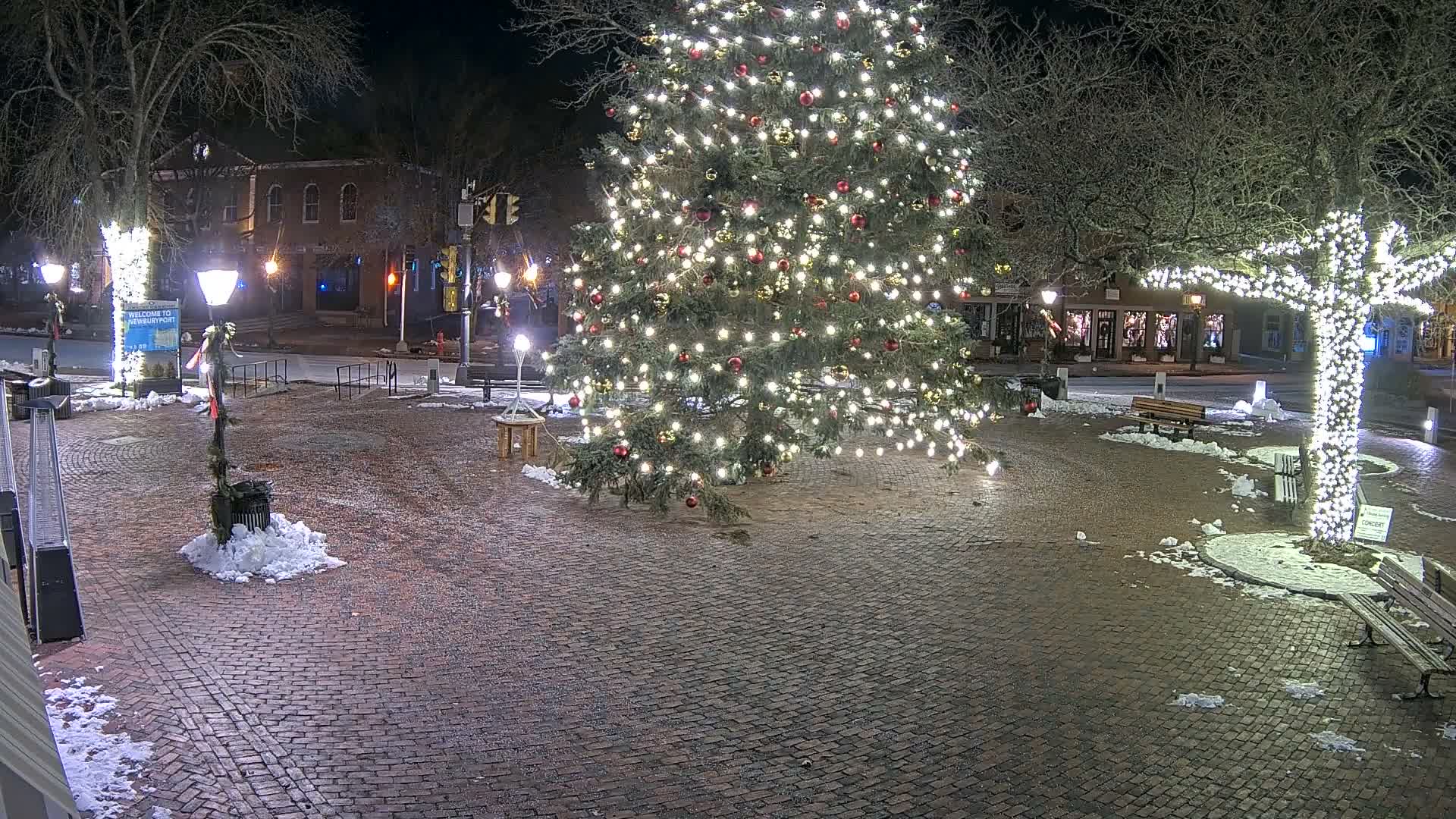 A festive town square is illuminated at night by a large, brightly decorated Christmas tree and other trees wrapped in white lights, with patches of snow scattered across the brick pavement on a clear, cold evening.