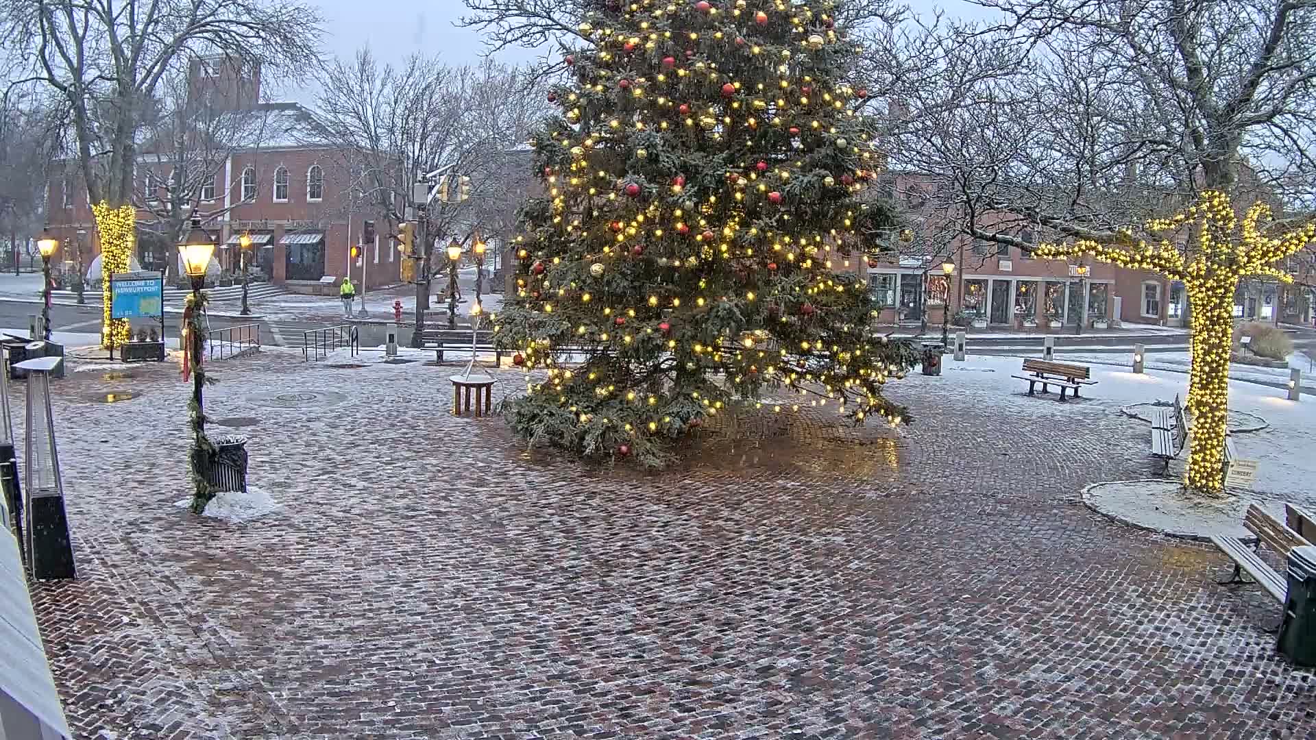 A festive town square is illuminated at night by a large, brightly decorated Christmas tree and other trees wrapped in white lights, with patches of snow scattered across the brick pavement on a clear, cold evening.
