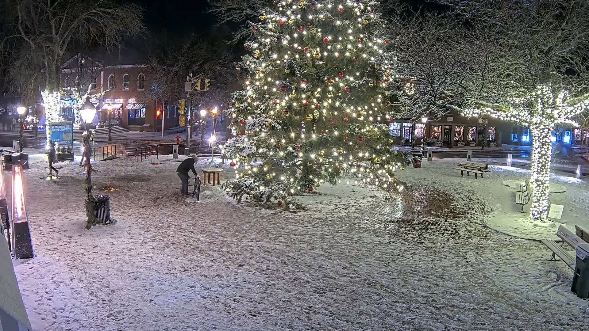 A festive town square is illuminated at night by a large, brightly decorated Christmas tree and other trees wrapped in white lights, with patches of snow scattered across the brick pavement on a clear, cold evening.