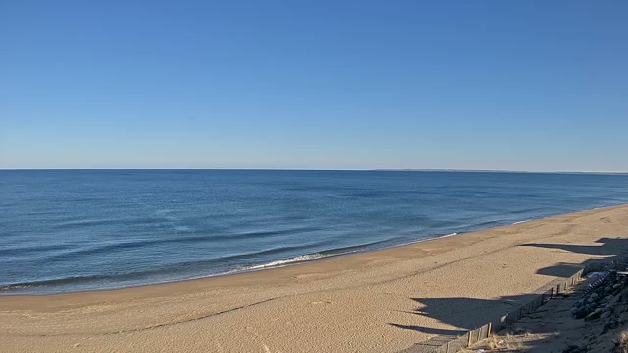 A wide sandy beach slopes down to a calm, blue ocean under a clear blue sky.