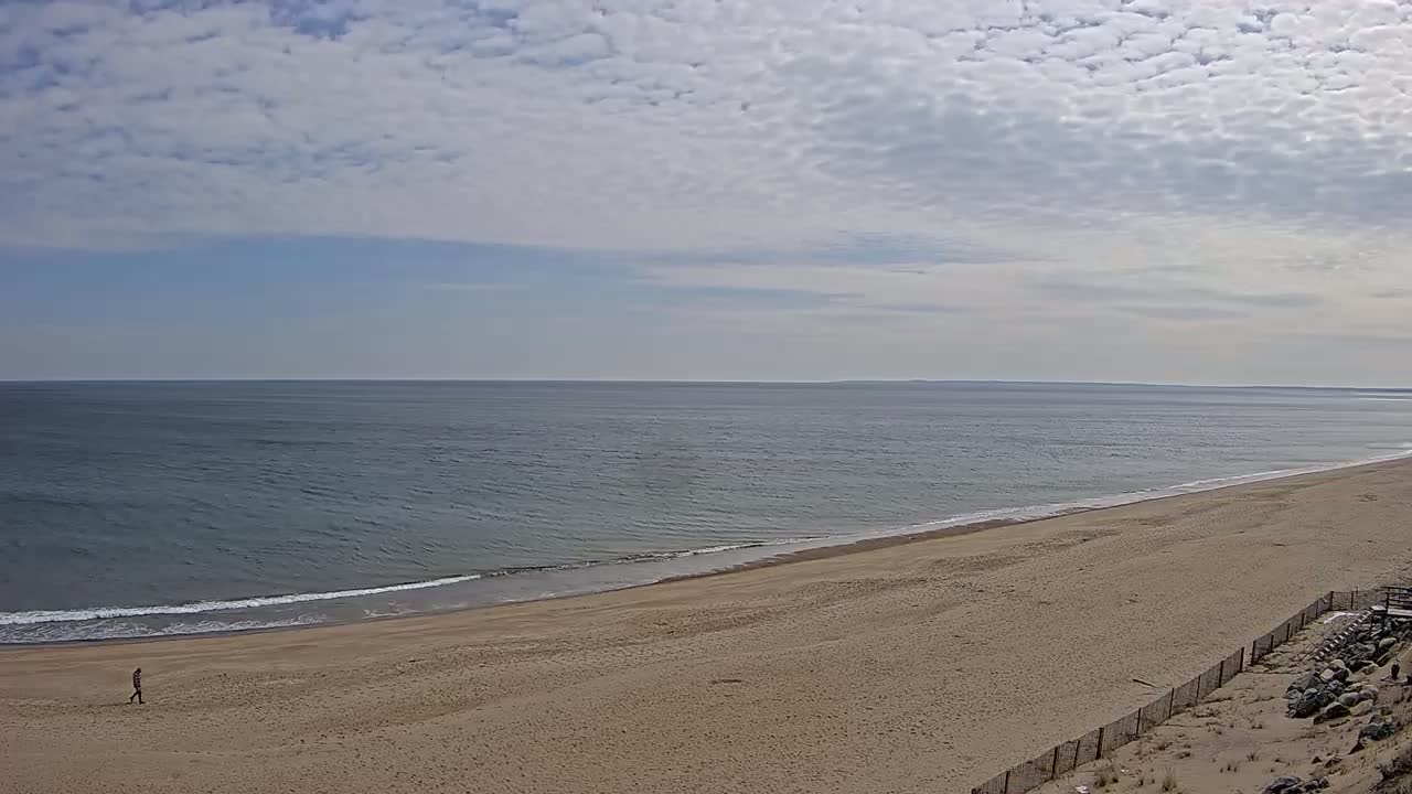 A person walks along a wide, sandy beach next to a calm ocean under a mostly cloudy sky.