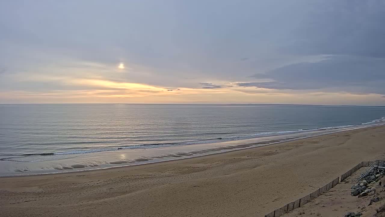 A sandy beach stretches along a calm ocean under a pale sunrise sky.