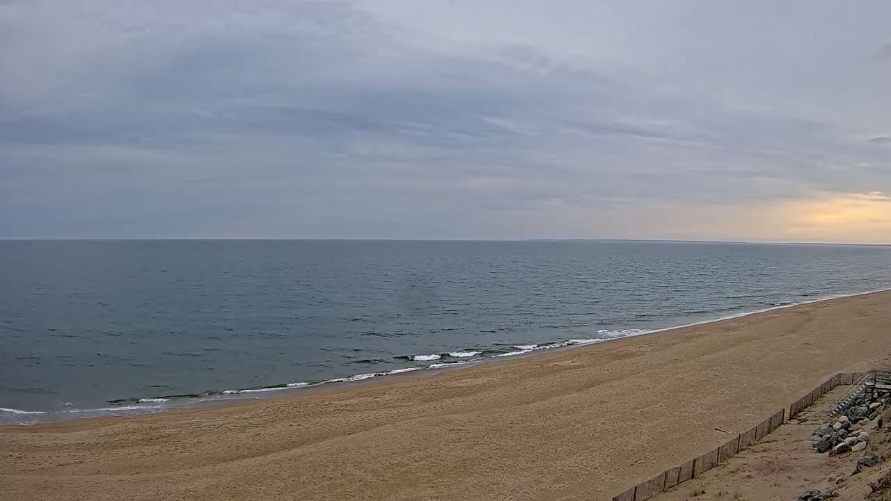 A long, sandy beach slopes gently down to a calm, gray-blue ocean under a mostly cloudy sky.