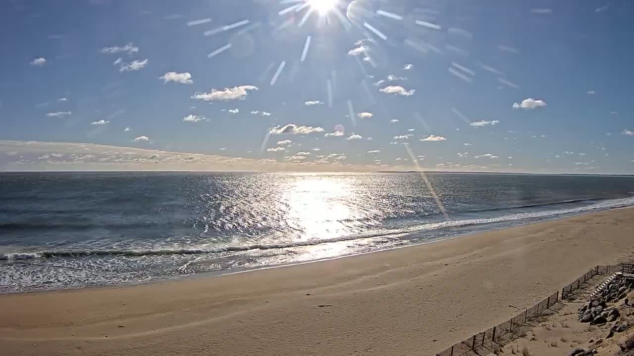 A sandy beach stretches toward a calm ocean under a bright, sunny sky with scattered clouds.