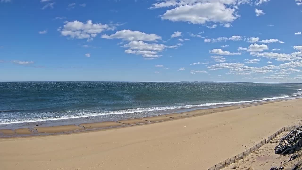 A wide sandy beach slopes gently down to a wavy ocean under a mostly sunny sky with scattered cumulus clouds.