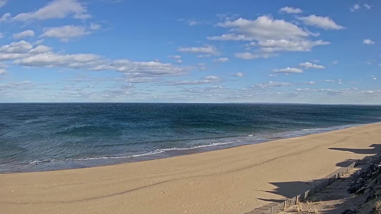 A sandy beach slopes down to a calm, blue ocean under a mostly sunny sky with scattered clouds.