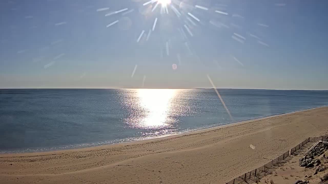 A sandy beach slopes down to a calm ocean reflecting the bright sunlight on a clear day.
