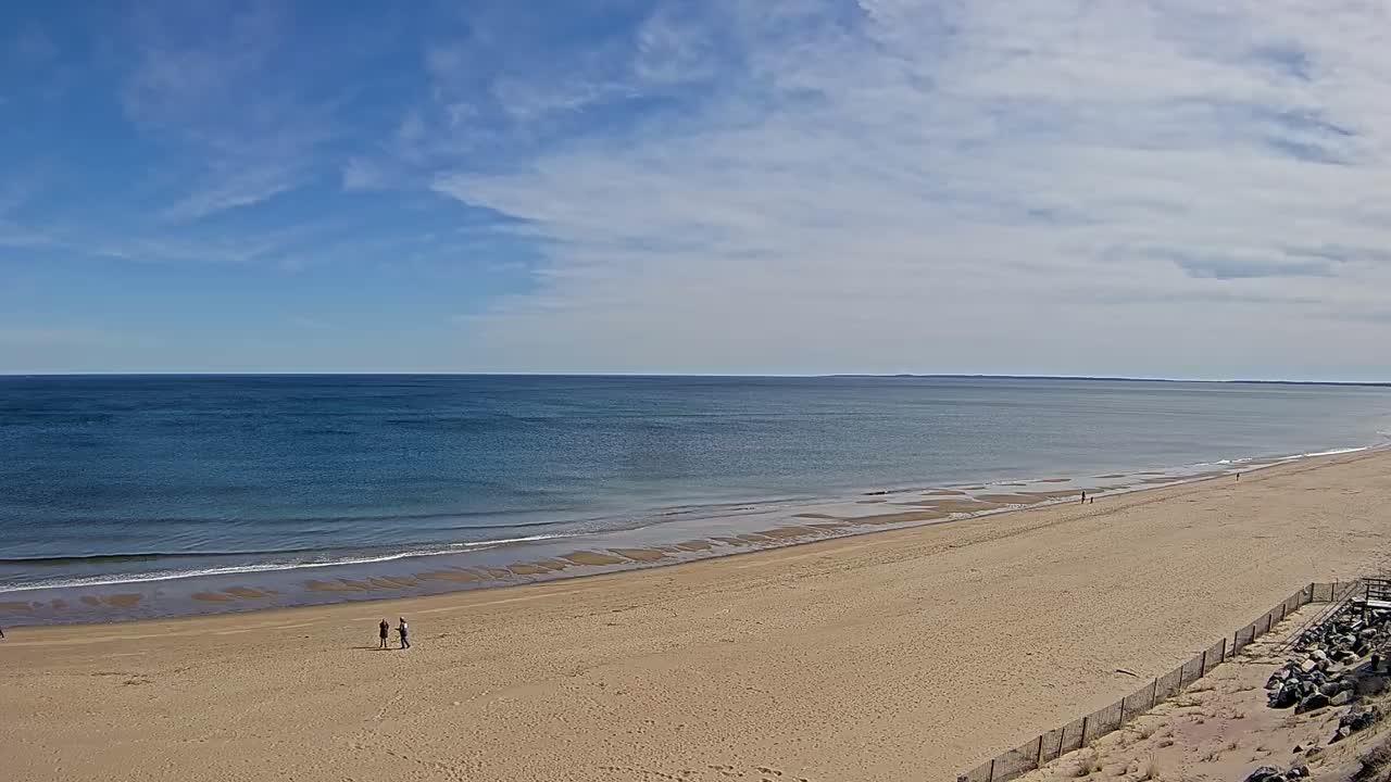 A long sandy beach stretches towards a calm ocean under a mostly sunny sky with some scattered clouds.