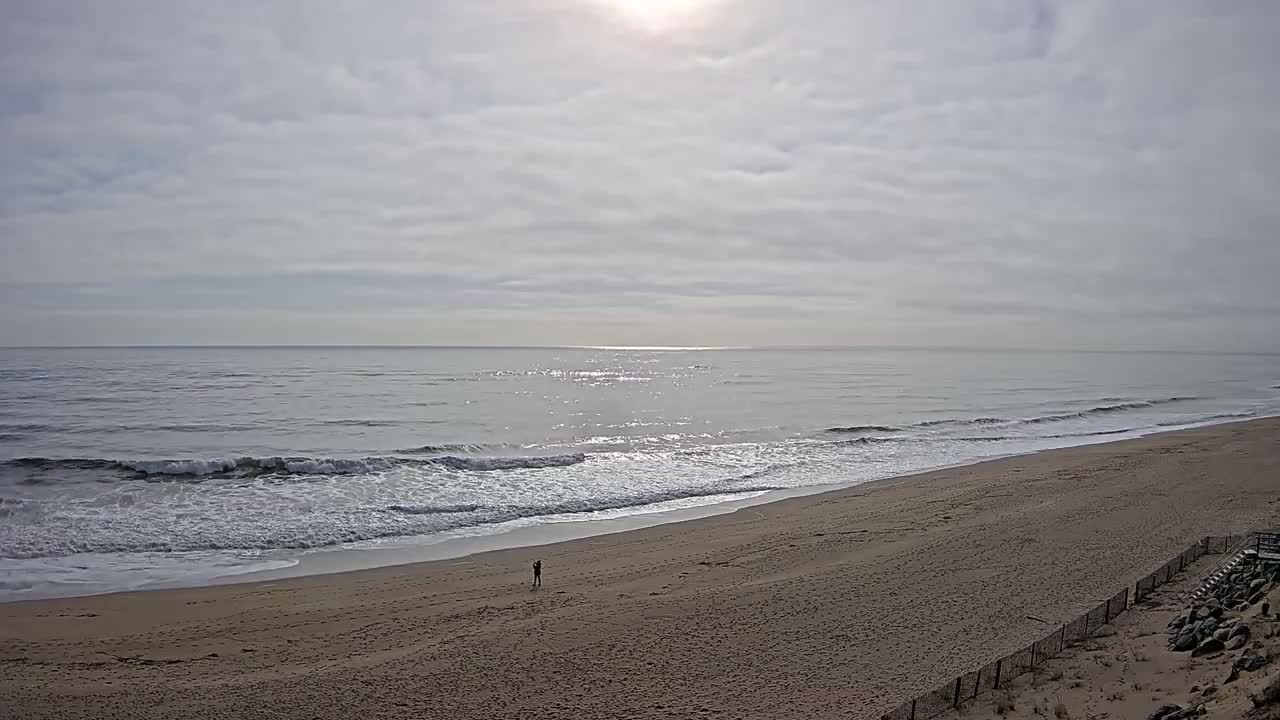 A person stands on a sandy beach with gentle waves rolling in under a mostly cloudy sky.