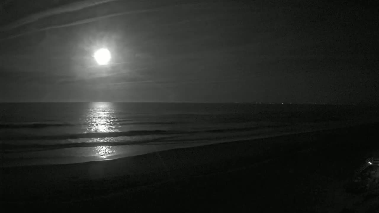A bright moon illuminates a dark beach and gently rolling ocean waves at night under a mostly clear sky.