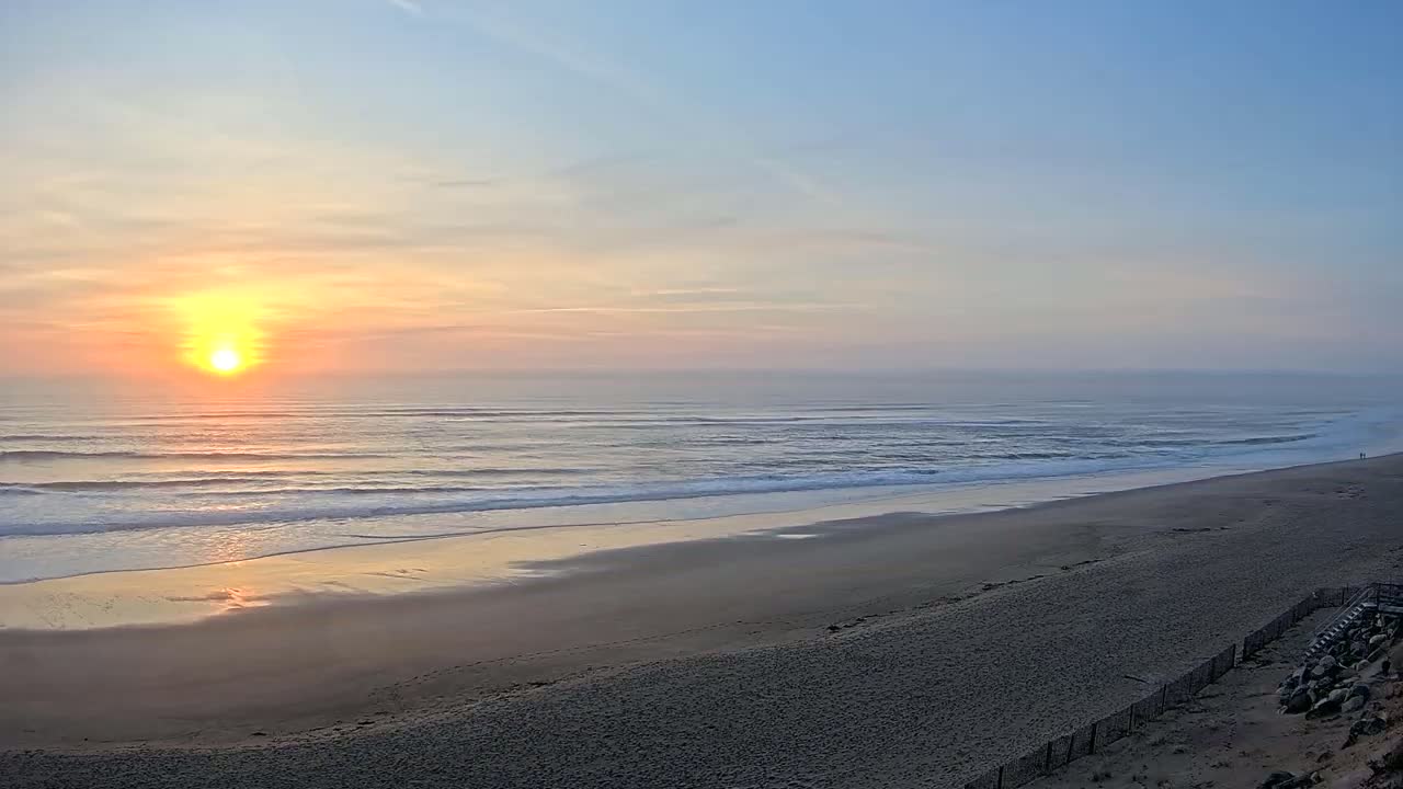 A hazy sunrise illuminates a wide, sandy beach and gently rolling ocean waves.