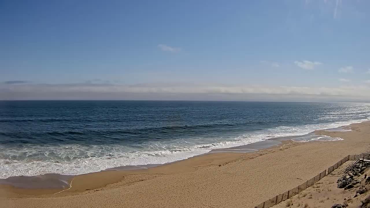 A sandy beach with gentle waves rolls onto the shore under a mostly clear, sunny sky.