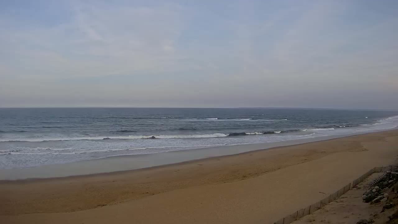 A long, sandy beach slopes gently down to a calm ocean with small waves under an overcast sky.