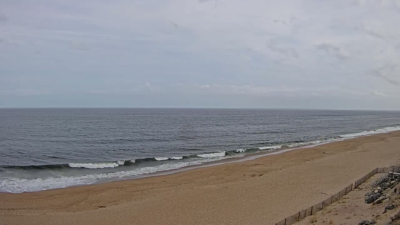 A wide sandy beach slopes down to a dark blue ocean with small waves under a mostly cloudy sky.