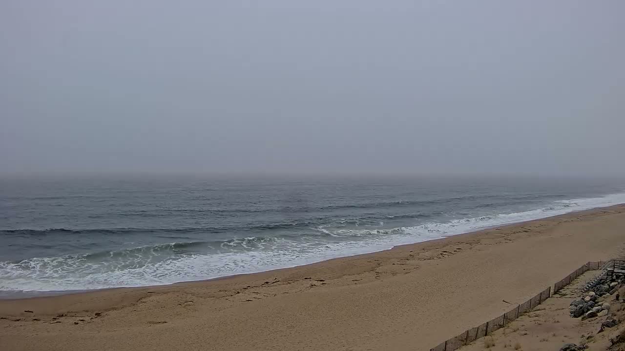 A foggy, overcast day reveals a sandy beach with gently rolling waves meeting the shore.