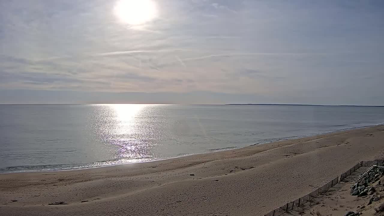 A sandy beach slopes down to a calm ocean reflecting the bright sunlight under a mostly clear sky.