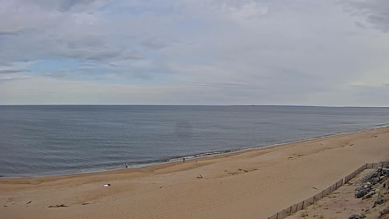 A wide, sandy beach stretches along a calm, gray ocean under a mostly cloudy sky.