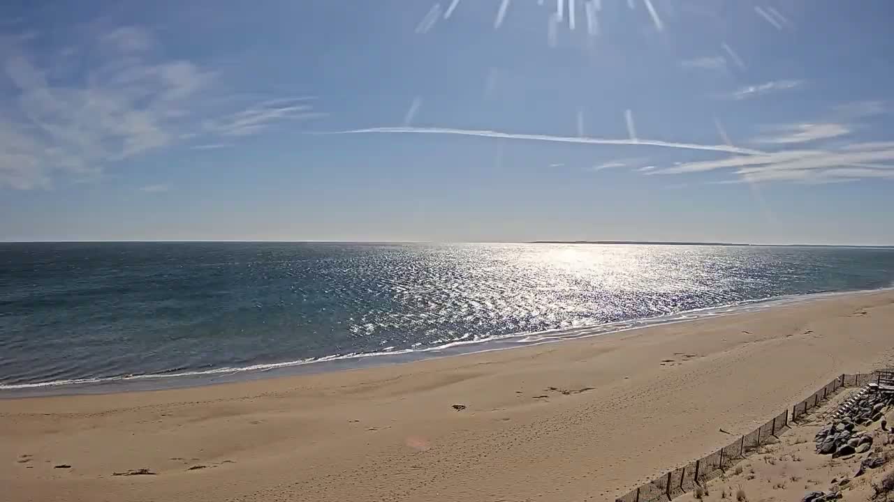 A sandy beach stretches out to a calm, sunlit ocean under a mostly clear, bright blue sky.