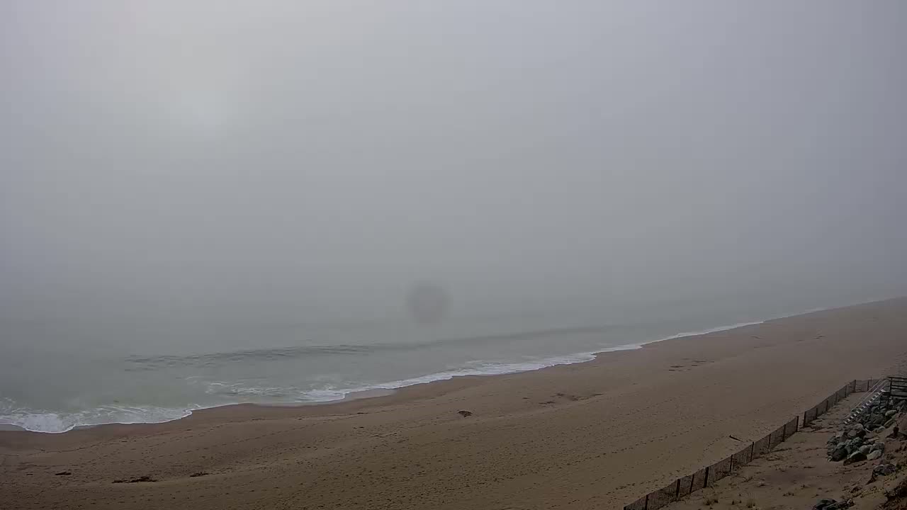 A foggy beach scene shows a sandy shore meeting a gray ocean with gentle waves, partially obscured by dense fog.