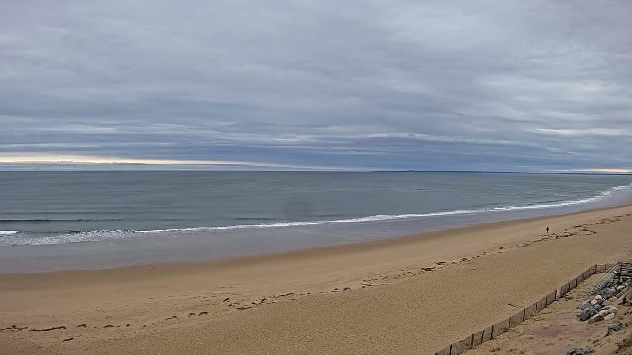 A long, sandy beach stretches towards a calm, gray ocean under a cloudy sky.