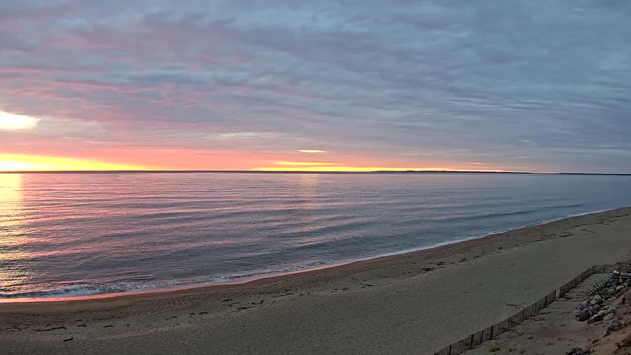 A sandy beach slopes down to a calm ocean under a sunrise sky with pink and purple clouds.