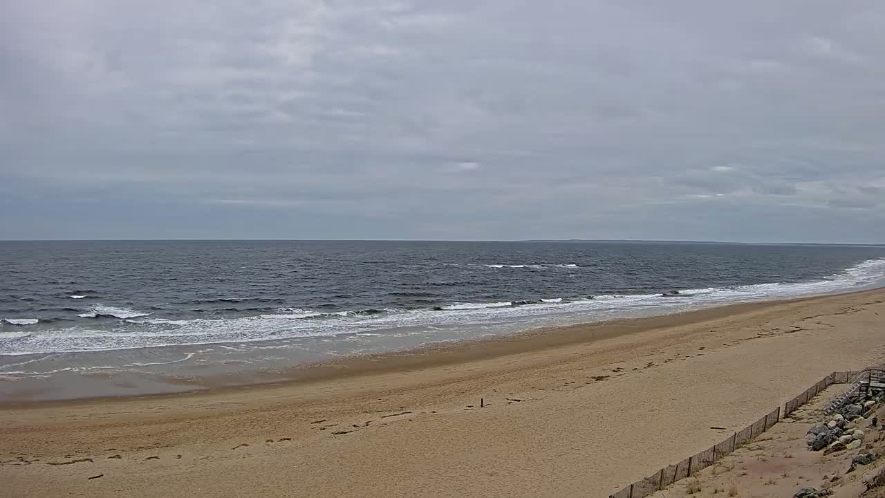 A wide sandy beach slopes down to a dark-blue ocean with small waves under a cloudy sky.