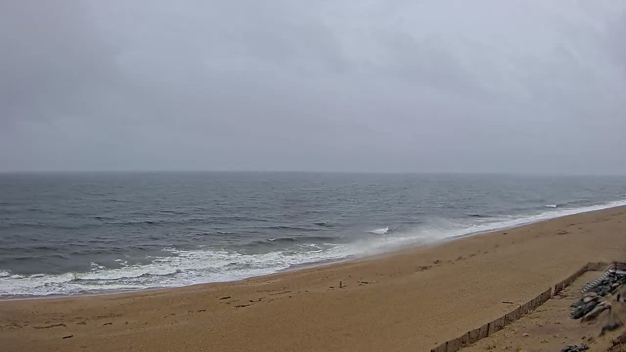 A sandy beach with moderate waves meets a gray, overcast ocean under a cloudy sky.
