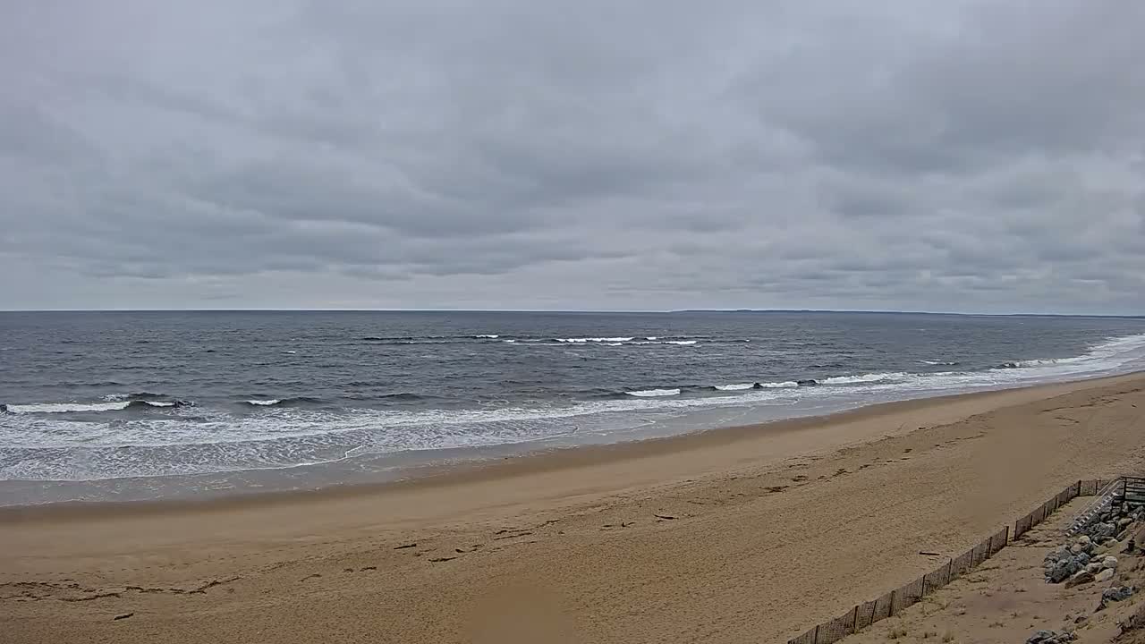 A wide sandy beach slopes down to a dark gray ocean with small waves under a cloudy sky.
