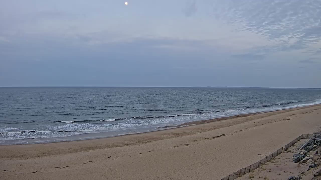 A mostly clear, daytime sky with a visible moon overlooks a wide sandy beach and gently waving ocean.